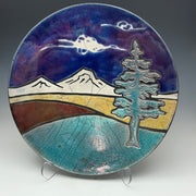 a round pottery plate with mountains and a tree on it